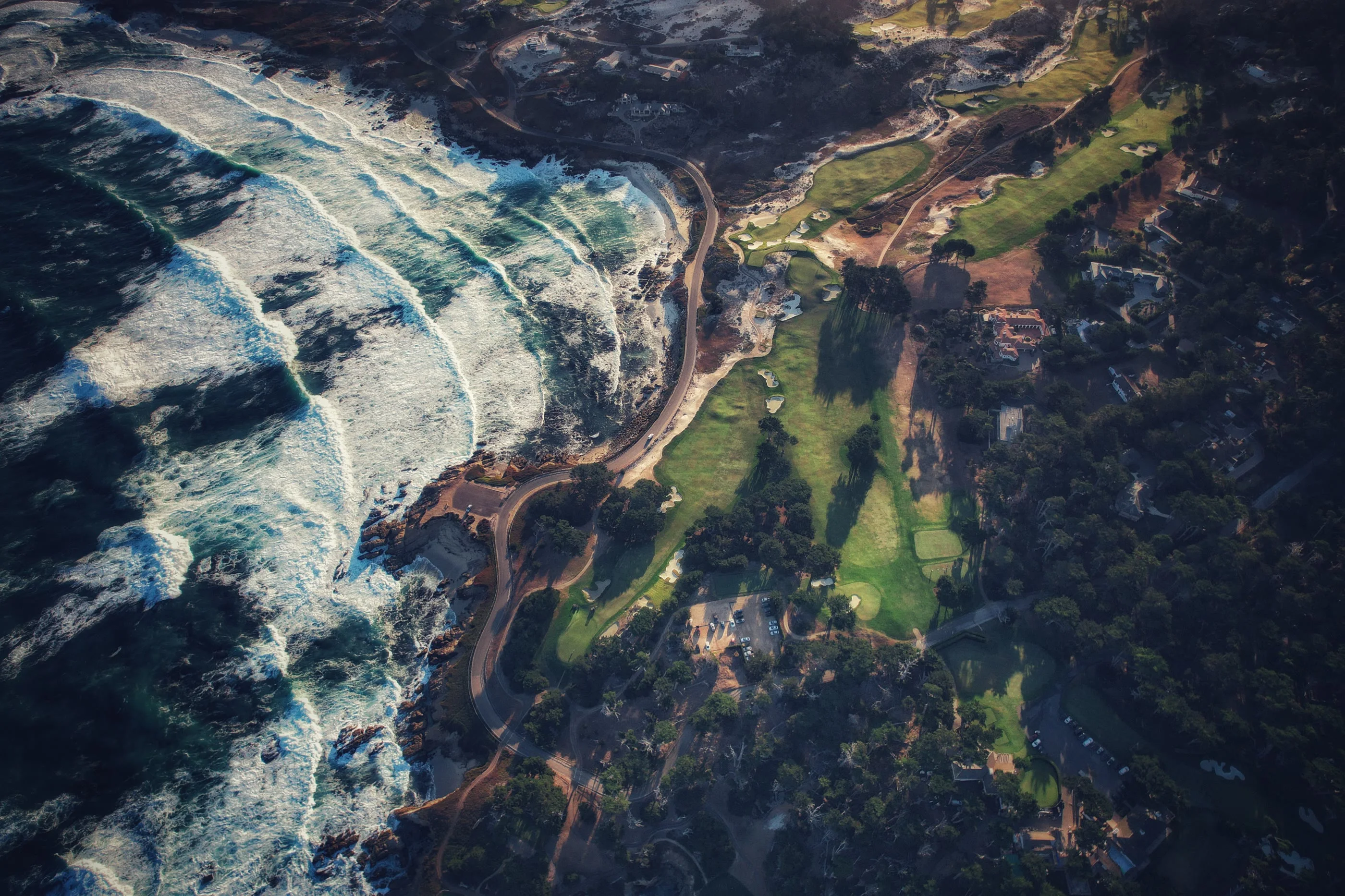 Aerial of the Monterey coast: surf breaking along the shore road, fairways running through the dunes